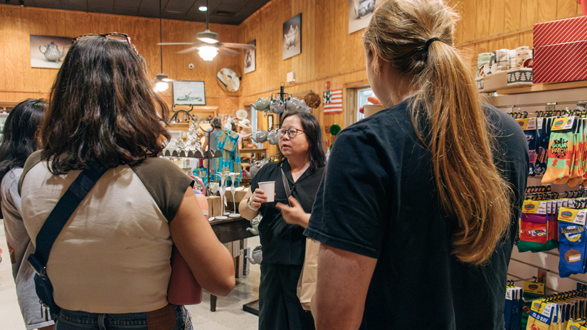 Dark haired woman with glasses holding a cup of tea and speaking to a group of female students.