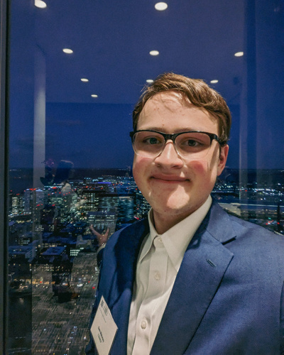 Young man in blue suit smiling with the night sky and a cityscape in the background.