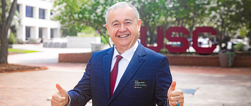 Fevzi Okumus, director of the University of South Carolina's School of Hospitality and Tourism Management, smiles and gives two thumbs up while standing on the USC campus with the university's iconic "USC" letters in the background.