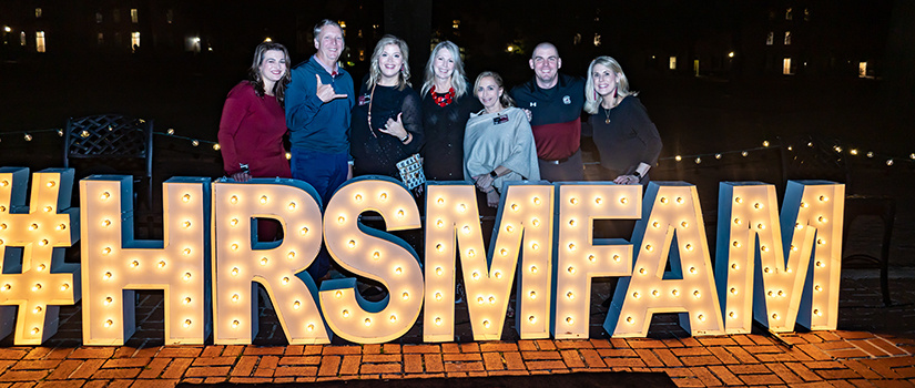 Seven people in attendance for the 2022 HRSM Homecoming Party pose for a photo behind large letters spelling #HRSMFAM.