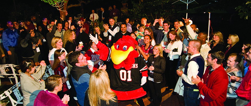HRSM guests, laughing and cheering, gather around a dancing Cocky while celebrating on the patio of McCutchen House during the HRSM Homecoming Party.