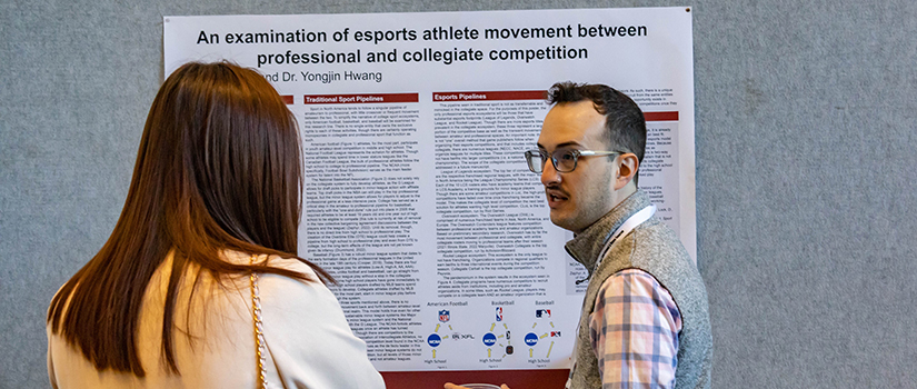 A Ph.D. student researcher stands with his poster presentation at the Sport Entertainment and Venues Tomorrow conference.