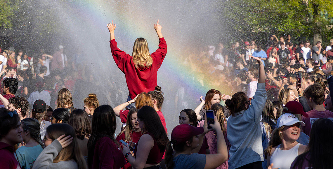 A large group of students gathered at the fountain in front of the Cooper Library. A rainbow can be seen in the water.