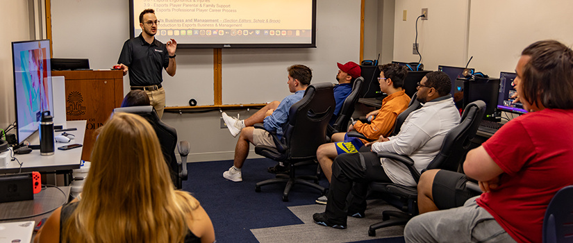 An instructor stands at the front of the esports laboratory giving a lecture to students.
