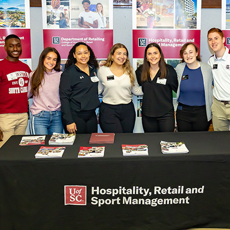 Seven HRSM students pose for a photo together behind a table while working Admitted Student Day.