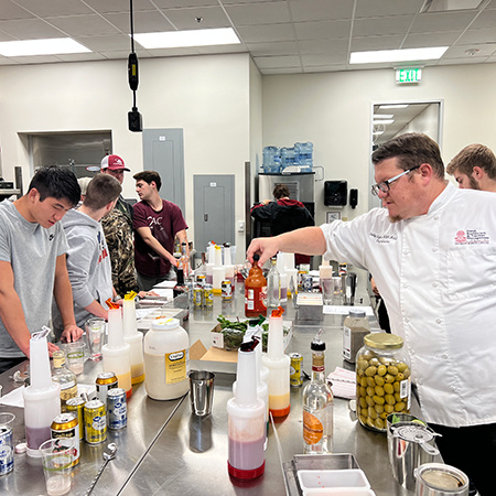 A group of students stand around a table in the Marriott Culinary Lab learning the process of how drinks are made in a restaurant operation.
