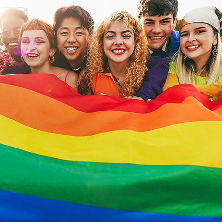Six young adults smile with a large pride flag in front