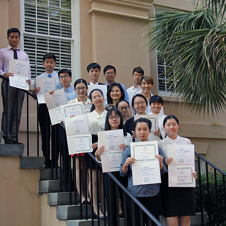 A group of visiting international students hold up their certificates of completion while standing on the steps of the McCutchen House.