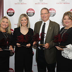 Alumni Award winners stand holding their awards in front of the HRSM Alumni Society step and repeat banner