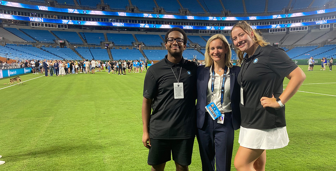 Two Tepper Scholars pose for a photo with a member of the Carolina Panthers' staff on the field at Bank of America Stadium.