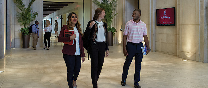 This is a photograph of students talking and walking through the law school lobby carrying books. In the foreground, there are two female students on the left and one male student on the right, and  there are two other students standing in the background talking and another standing farther behind in the distance.