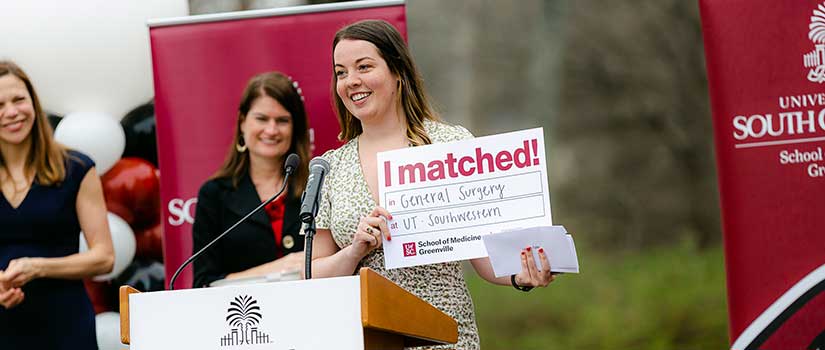 Female Med student holds an "I Matched" sign while standing at a podium
