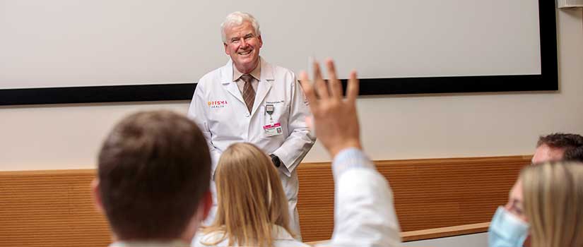 Medical student raises his hand in class with professor at the front of the room.