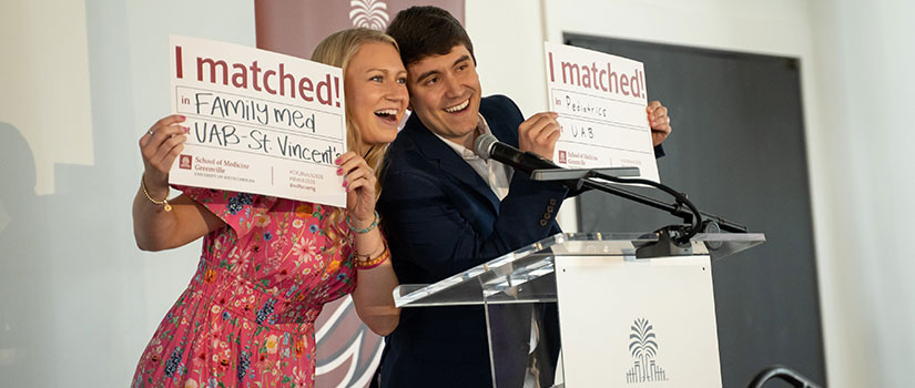 Female Med student holds an "I Matched" sign while standing at a podium