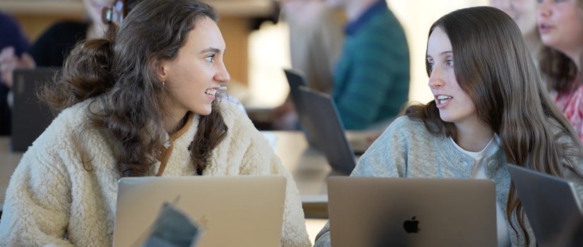 two female students talking to each other; laptops on the table and open