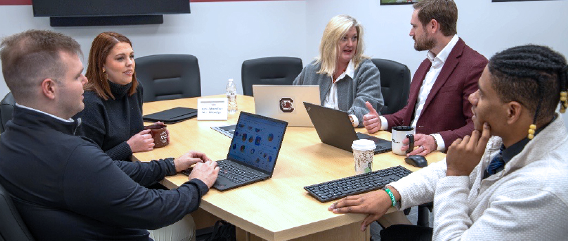five people sitting at a table talking and working on laptops