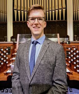 Thomas Russell stands in front of a pipe organ wearing a grey blazer, blue shirt and tie. 