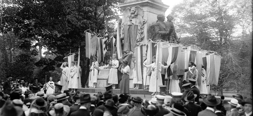 women with banners stand in front of a crowd near a monument in washington, DC, in 1918 to advocate for women's suffrage 