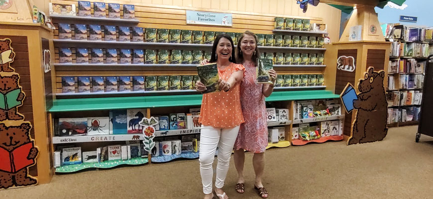 two women hold up books in front a large display case of books