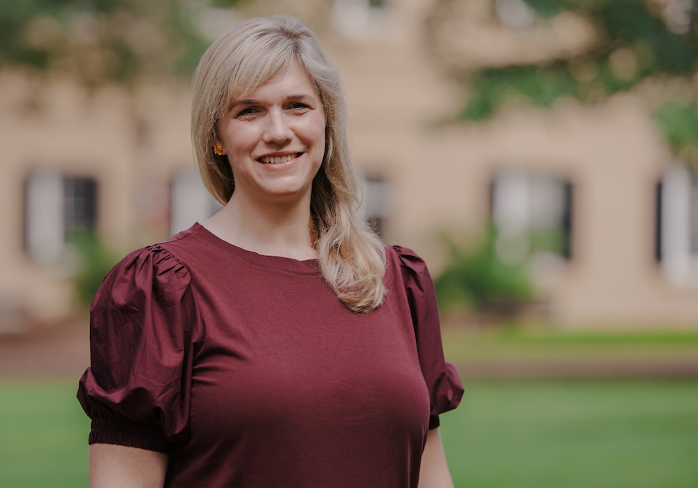 Blond-haired woman in garnet shirt smiles in front of beige building