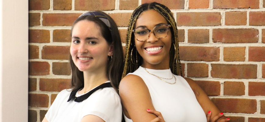 A pair of women, one with braided hair and glasses, the other with a headband, pose back-to-back, smiling in front of a brick wall.