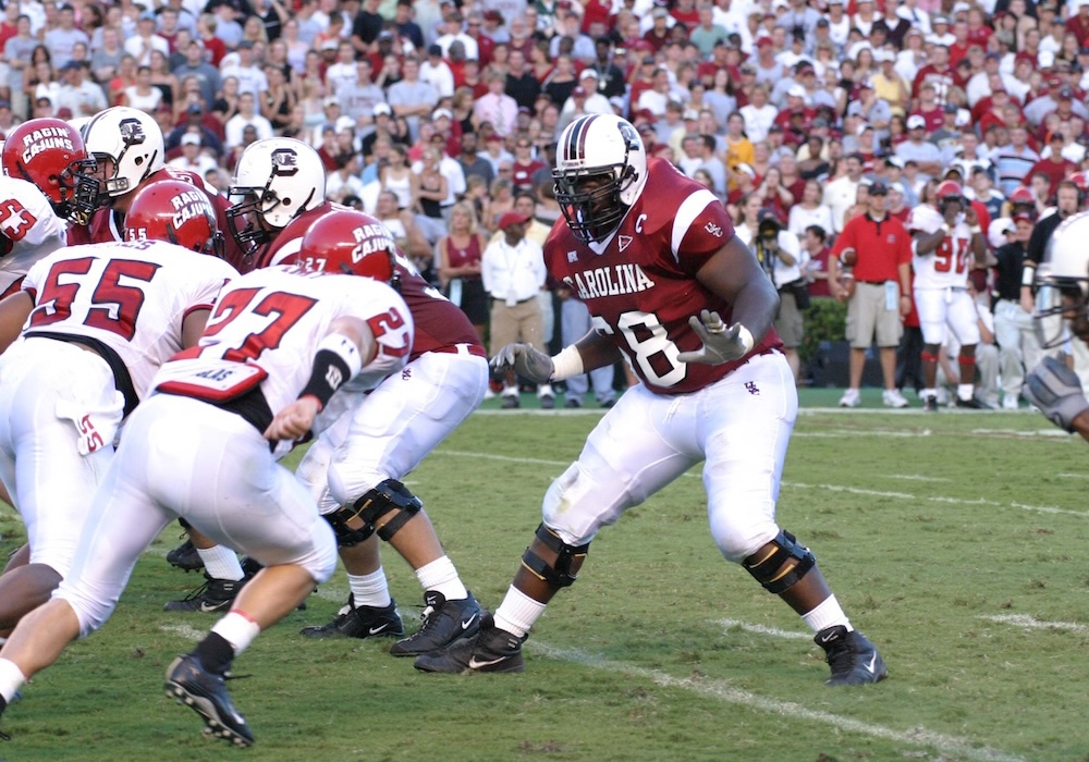 football player in white helmet, garnet jersey and white pants backpedals during a game