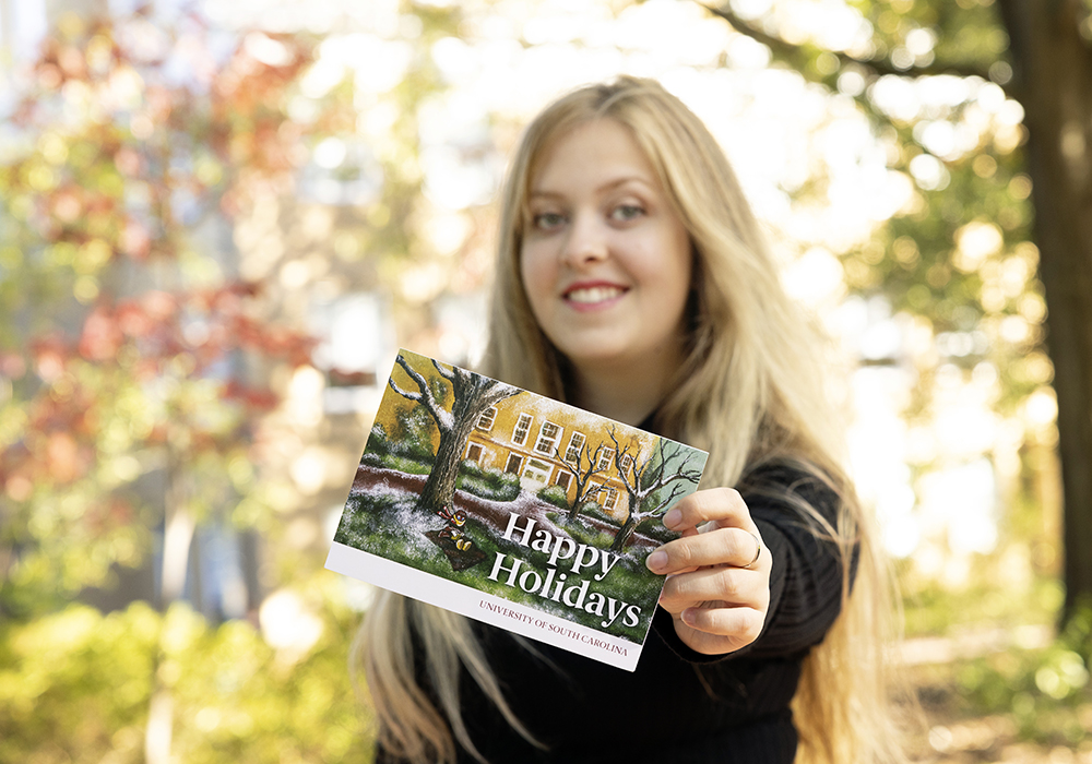 Lauren Puttock holds out a card featuring her painting of Cocky reading on campus in the snow.