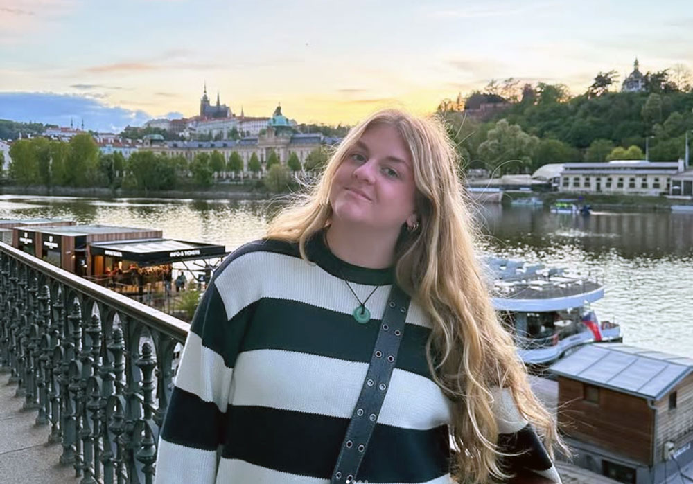 a woman stands in front of a river with a city in the background