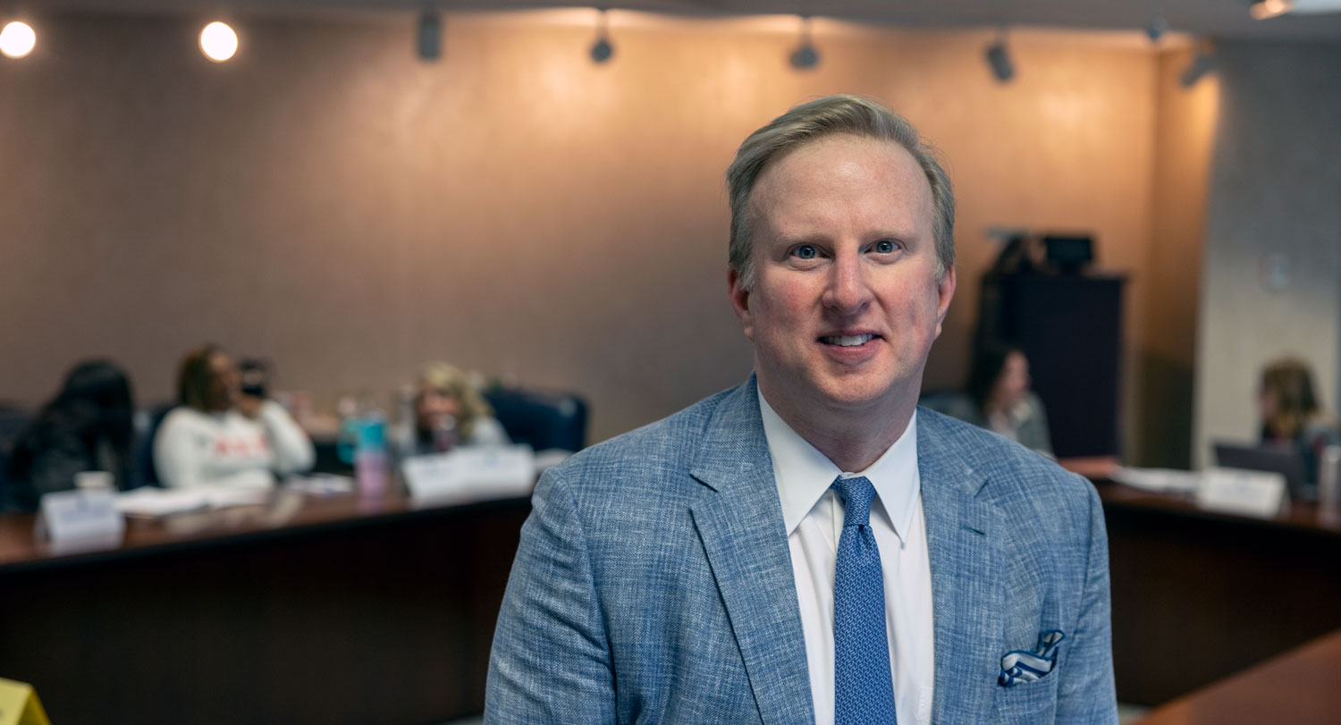 Man stands in front of a conference table with people in the background