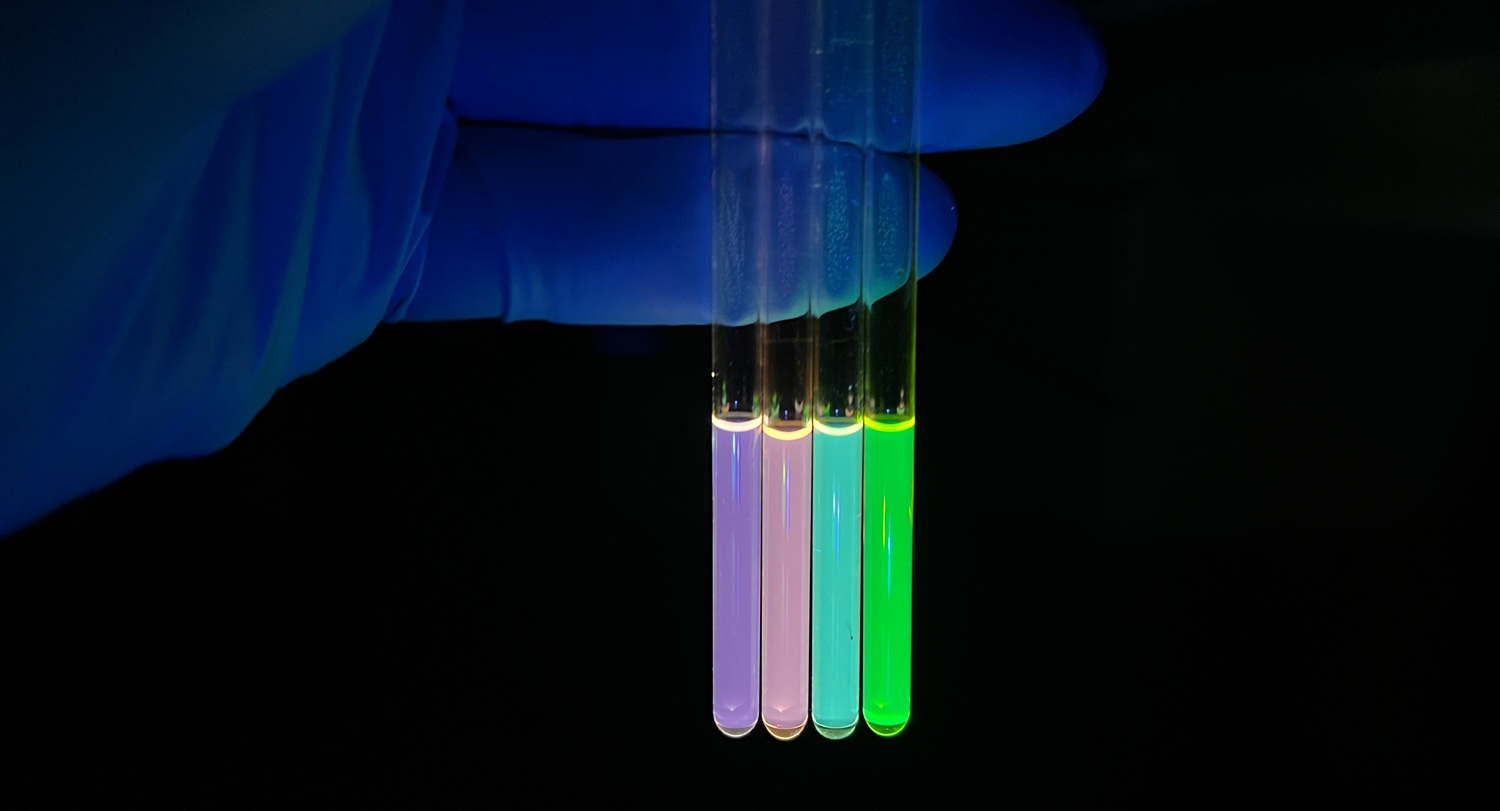 A gloved hand holds four tubes displaying fluorescent nanocyrstals in a dark space.