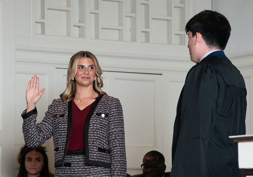 Woman stands with her right hand in the air looking at another student
