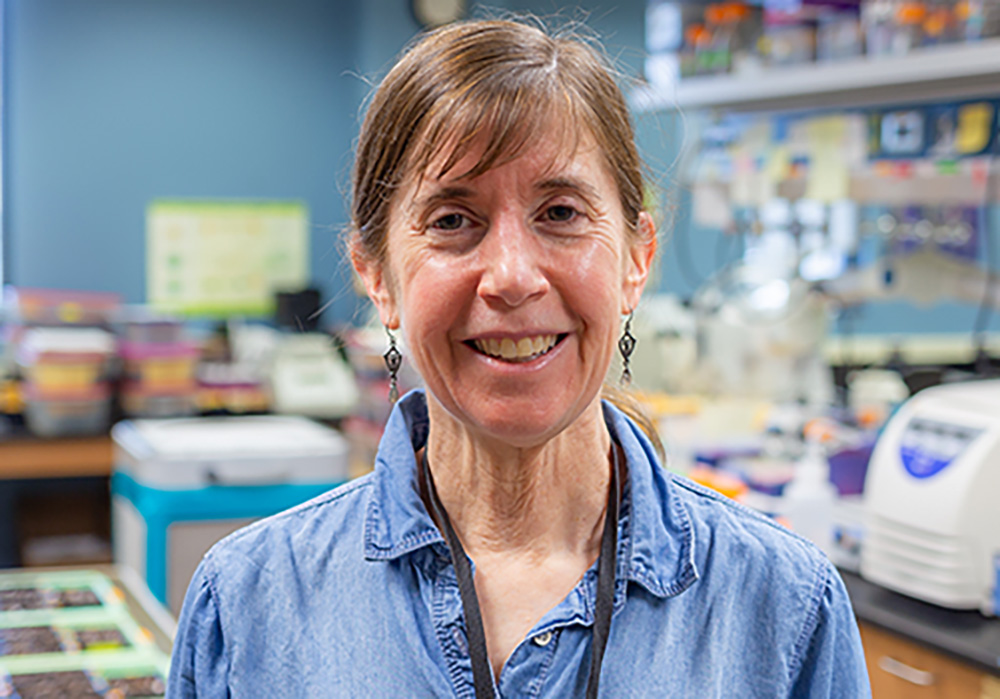 A white woman with brown hair tied back and wearing a blue button-down shirt smiles from a biology lab setting