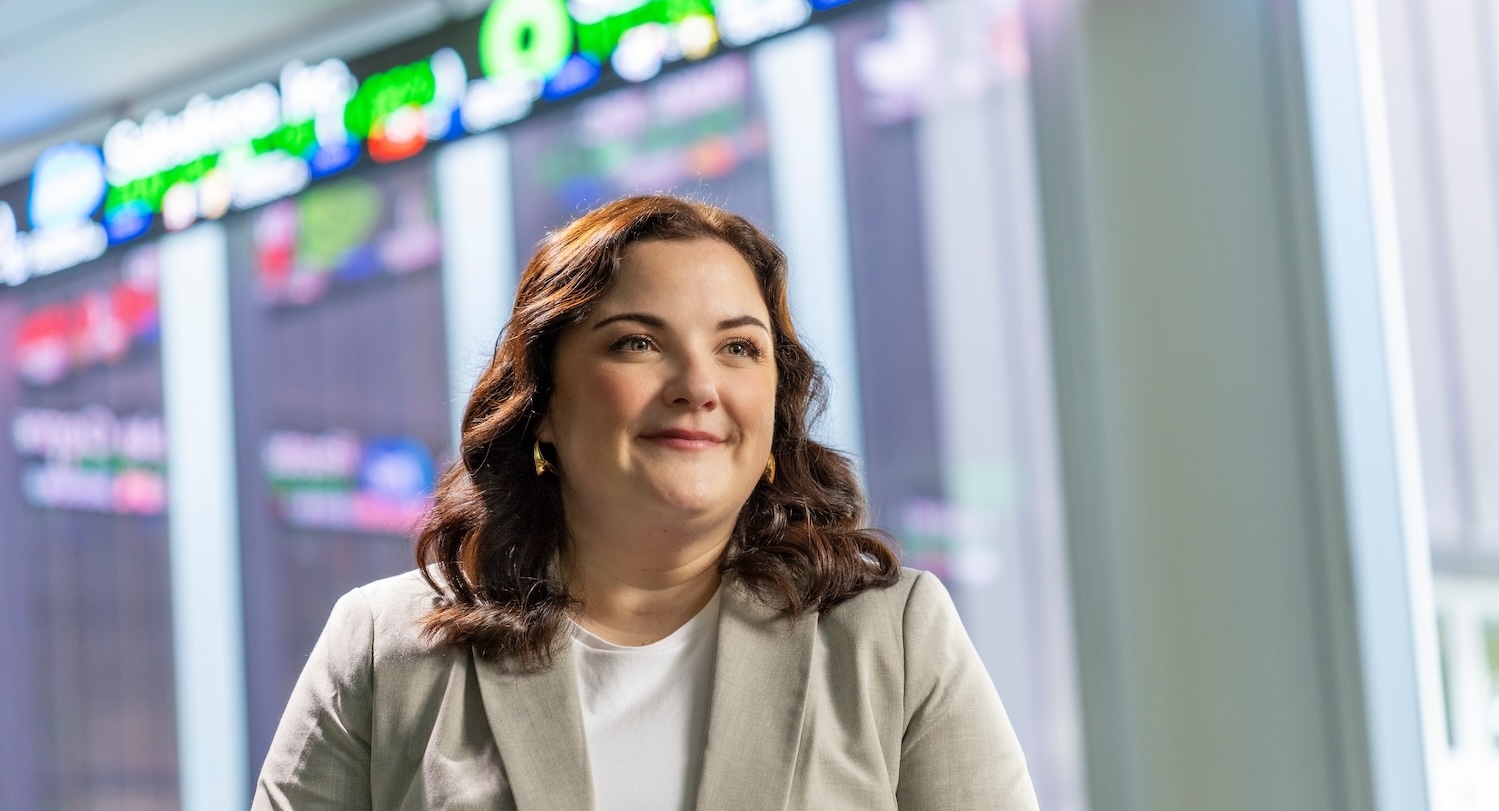 USC alumna Stephanie Kyle smiles warmly while standing in front of a financial data display board showing stock tickers.