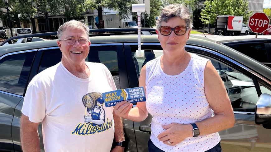 two people stand in front of a car holding a bumper sticker that says heat watch climate science in motion