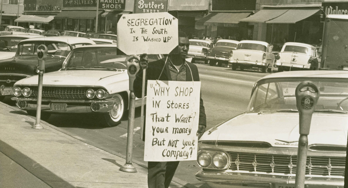 A man, possibly Gene Hawkins with 2 protest signs walking down Main Street, in which is  lined with cars. The two protest signs read: Segregation in the south is washed up and Why shop in stores that want your money but not your company? Many business and signs can be seen in the background.