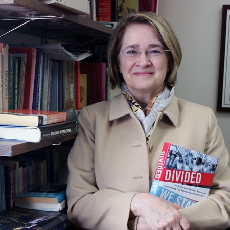historian marjorie spruill stands in front of a bookcase holding a copy of her book Divided We Stand: The Battle Over Women's Rights and Family Values That Polarized American Politics 