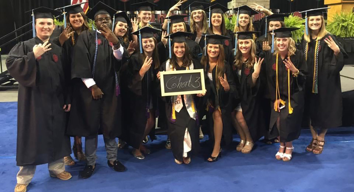 Group in graduation gowns holding sign reading "Cohort 3"