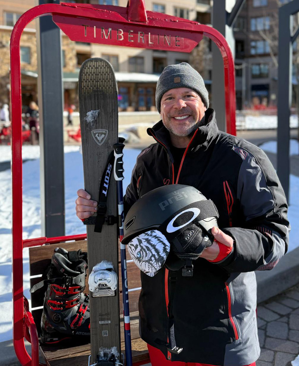 Man in winter jacket and knit hat holds skis and a helmet while standing beneath red Timberline ski lift sign at snowy mountain resort. 