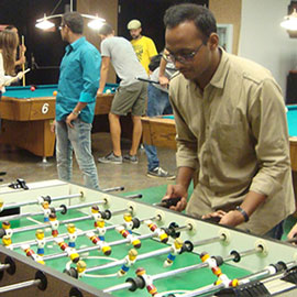 Students hanging out in the Golden Spur Game room. In the front of the photo several students are playing foosball and in the background there are students playing pool.