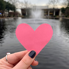 A hand holding pink heart paper cutout in front of the Thomas Cooper Library and fountain