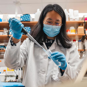 person with dark hair in lab wearing lab coat, blue mask and blue gloves