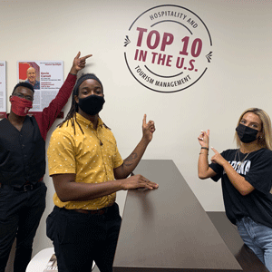 Three students wearing masks in front of a counter pointing to a wall badge that says top 10