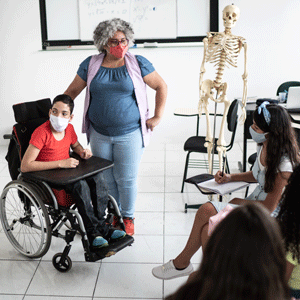 A teacher stands beside student using a wheelchair while other students sit in desks