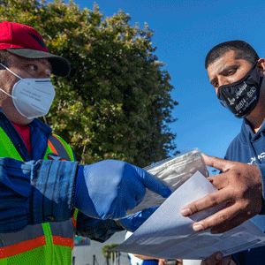 Man in red cap wearing gloves gives another man a packet of face masks
