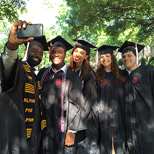Five students in caps in gowns take a selfie, the man on the left holds the camera