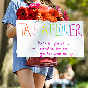 Student holding a bucket of flowers with a sign that says take a flower.