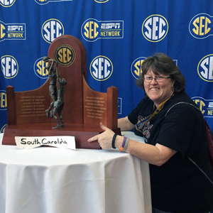 Woman stands next to the SEC women's basketball trophy with a handwritten south carolina sign in front and a backdrop with ESPN SEC Network logo on it