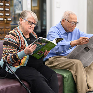 A man and a woman sit in a library, reading a magazine and newspaper.