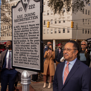 A man stands beside a historical marker documenting the enrollment of the first black student at USC during Reconstruction; people stand in the background