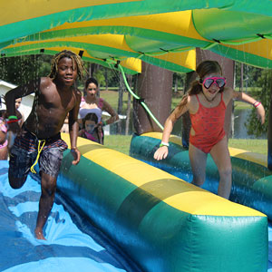 Two children in bathing suits smile as they run through an inflatable slip and slide.